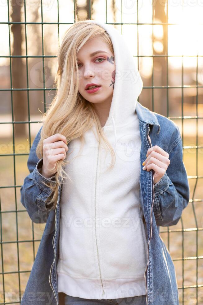 giovane donna con temporaneo dipinto fiori su il viso. adolescenziale ragazza con lungo biondo capelli indossare jeans giacca e felpa con cappuccio foto