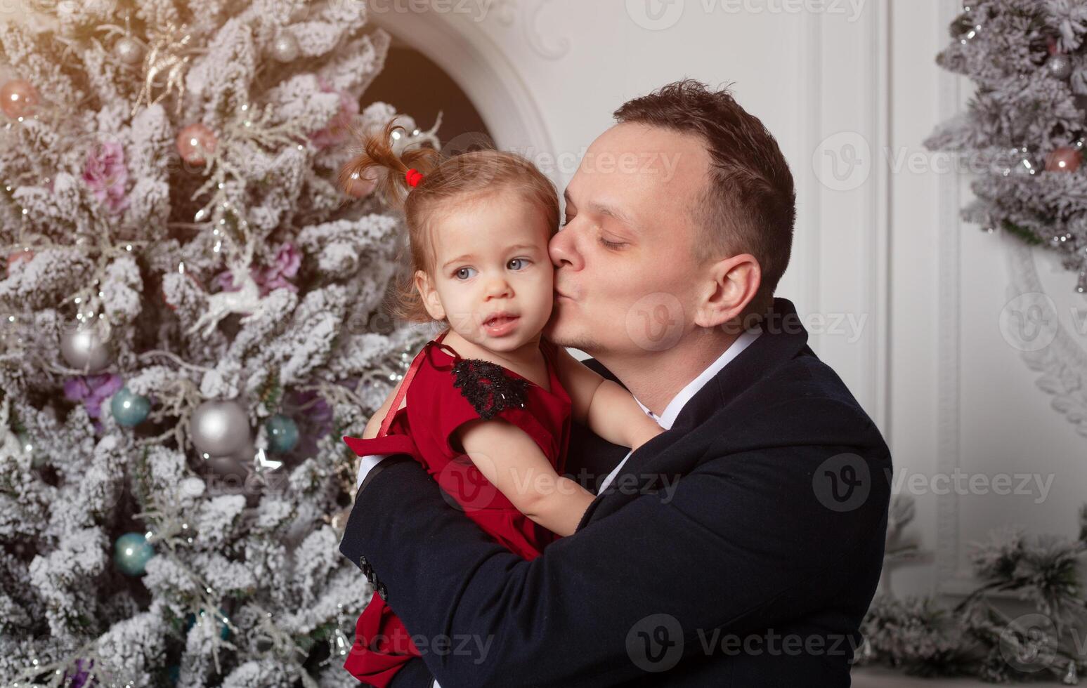 allegro Natale e contento nuovo anno. papà nel un' attività commerciale completo da uomo detiene il suo figlia vestito nel un elegante rosso vestito su il sfondo di il Natale albero foto