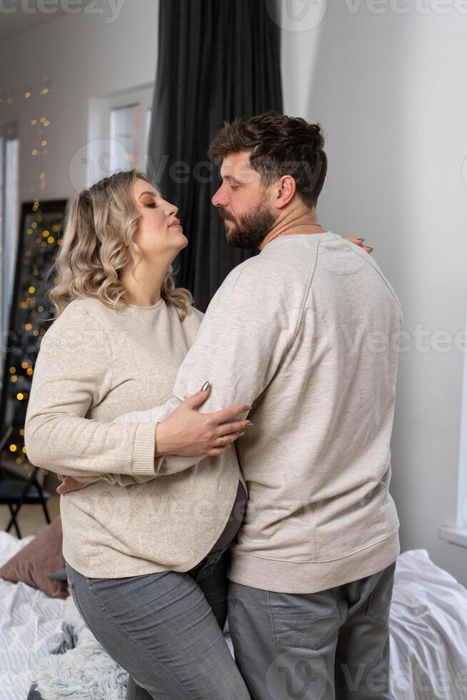 contento famiglia concetto. marito abbraccio pancia incinta moglie in piedi interno vivente camera vicino divano foto