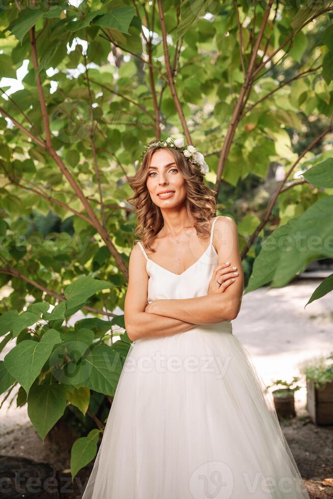 bellissimo ragazza nel bianca vestito con un' ghirlanda di fiori su sua testa è in piedi all'aperto nel il parco con verde foglia su il sfondo foto