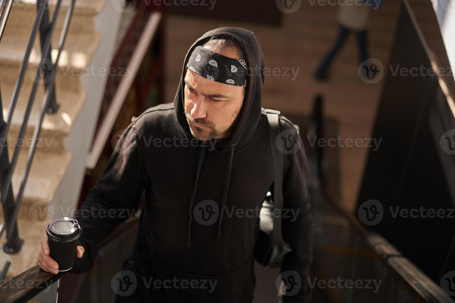 bello giovane con la barba lunga uomo nero felpa con cappuccio e bandana con caffè carta tazza va su il scala mobile. foto