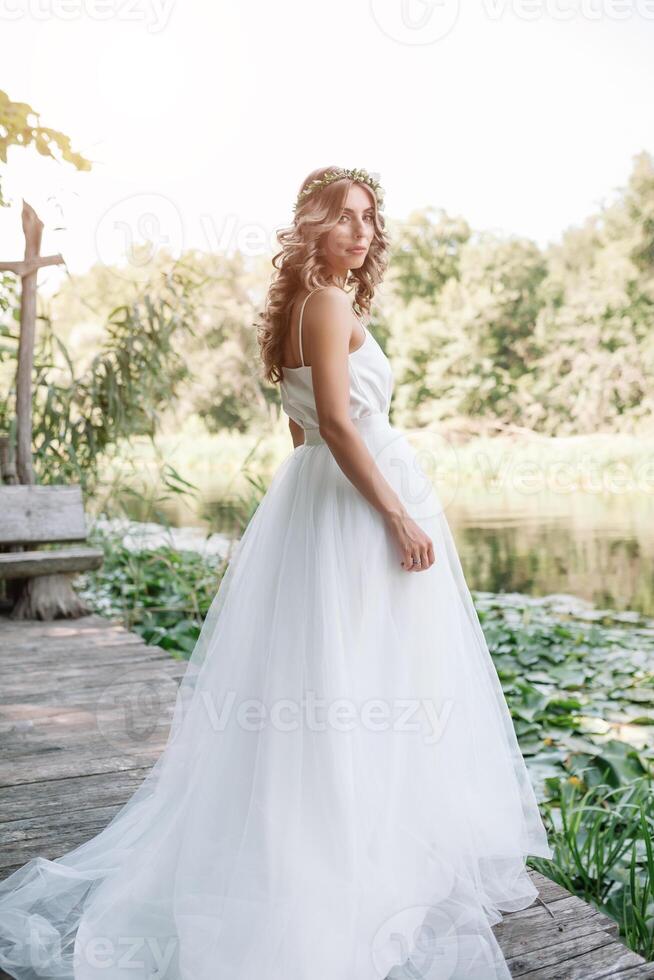 un' carino Riccio donna nel un' bianca nozze vestito con un' nozze mazzo e ghirlanda nel sua capelli in piedi indietro per il telecamera nel natura. concetto sfuggito sposa. inoltrare per un' contento luminosa futuro scappa foto