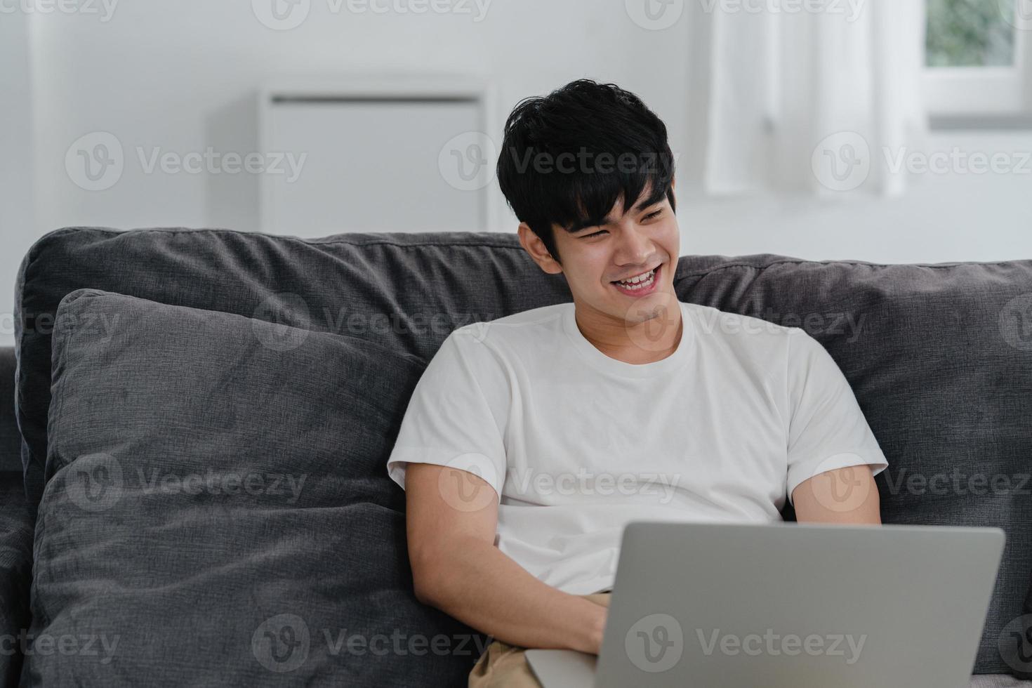 uomo asiatico freelance che lavora a casa, maschio creativo su laptop sul divano in soggiorno. giovane imprenditore proprietario di affari, gioca al computer, controlla i social media sul posto di lavoro al concetto di casa moderna. foto