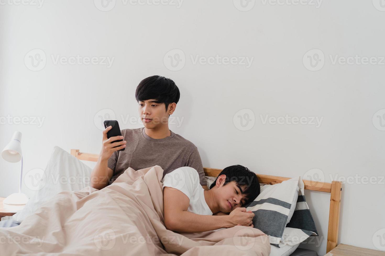 coppia gay asiatica utilizzando il telefono cellulare a casa. giovane asia lgbtq uomo felice relax riposo dopo il risveglio, controlla i social media mentre il suo ragazzo dorme sdraiato sul letto in camera da letto a casa al mattino concetto. foto