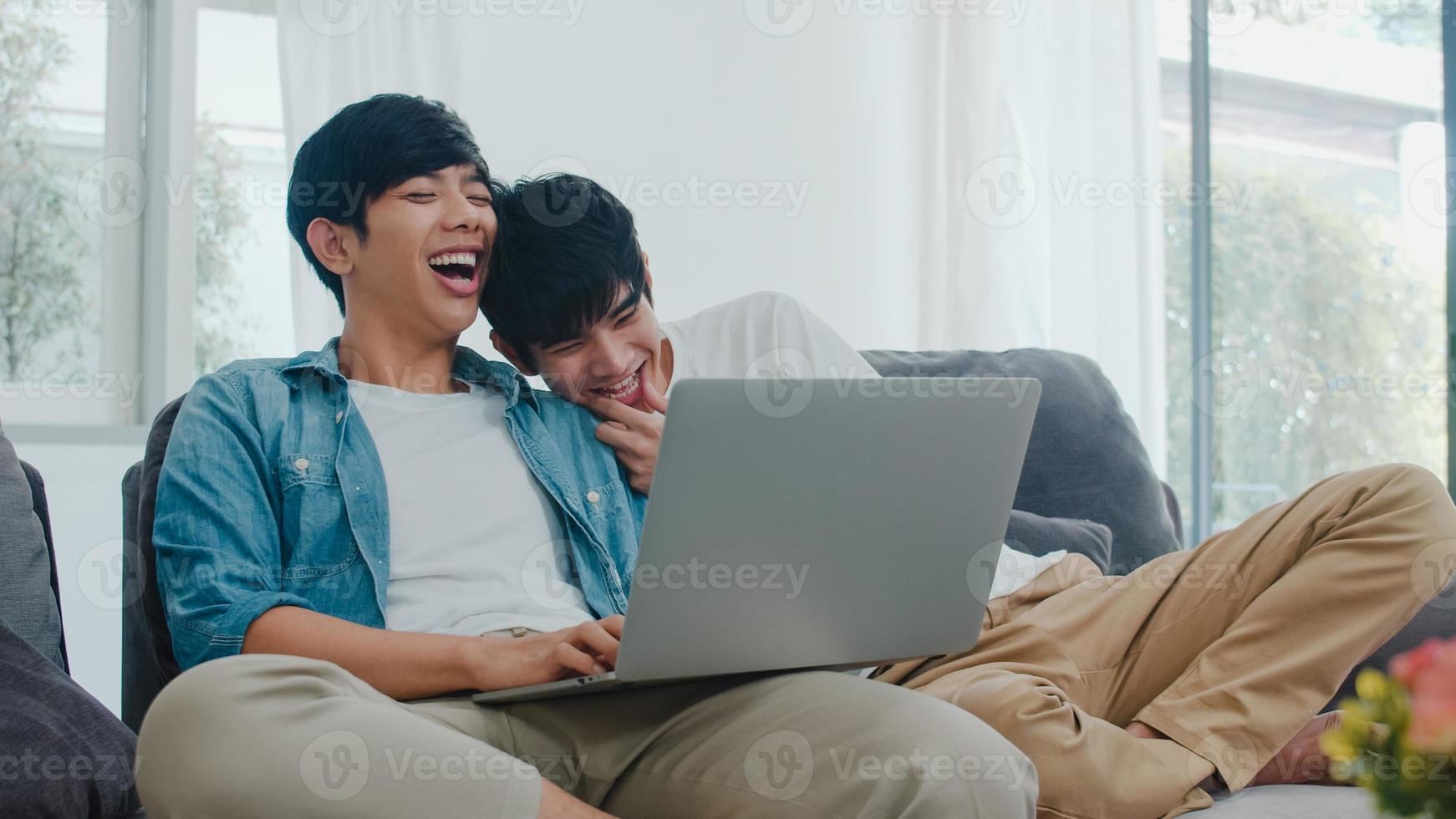 giovane coppia gay che utilizza computer portatile a casa moderna. gli uomini asiatici lgbtq si rilassano felici e si divertono usando la tecnologia guardando film in internet insieme mentre si trovano sul divano nel soggiorno al concetto di casa. foto