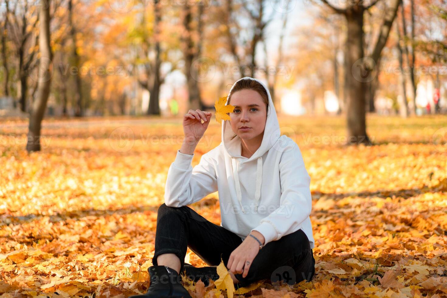 donna nel un' bianca maglione con un' cappuccio si siede su terra nel il parco e Tenere un' foglia nel sua mani foto