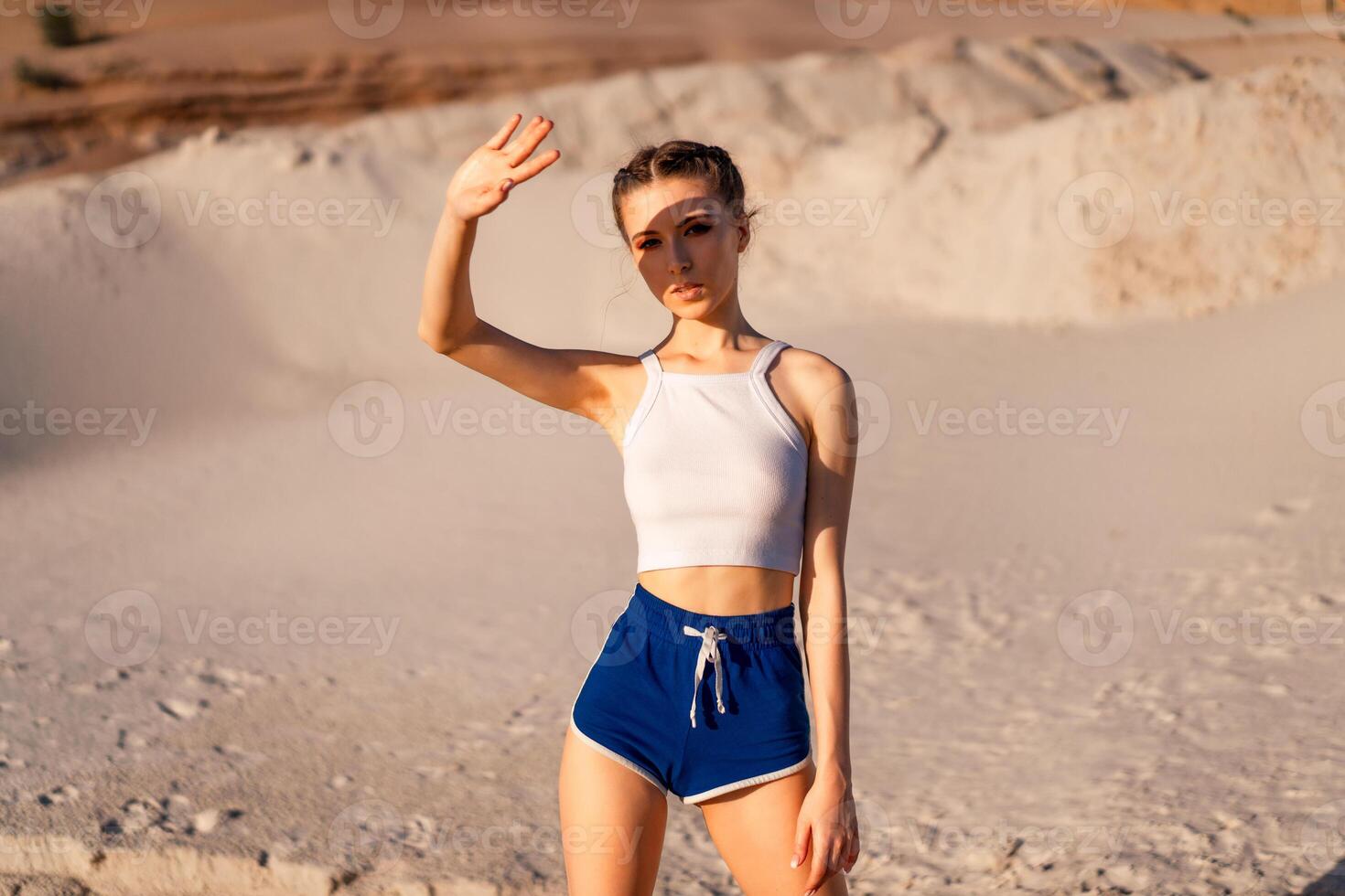 un' bellissimo ragazza nel un' sexy bianca superiore e corto pantaloncini sta nel il deserto e coperture sua occhi con il mano a partire dal il sole. un' mano ombra cascate su sua viso. foto