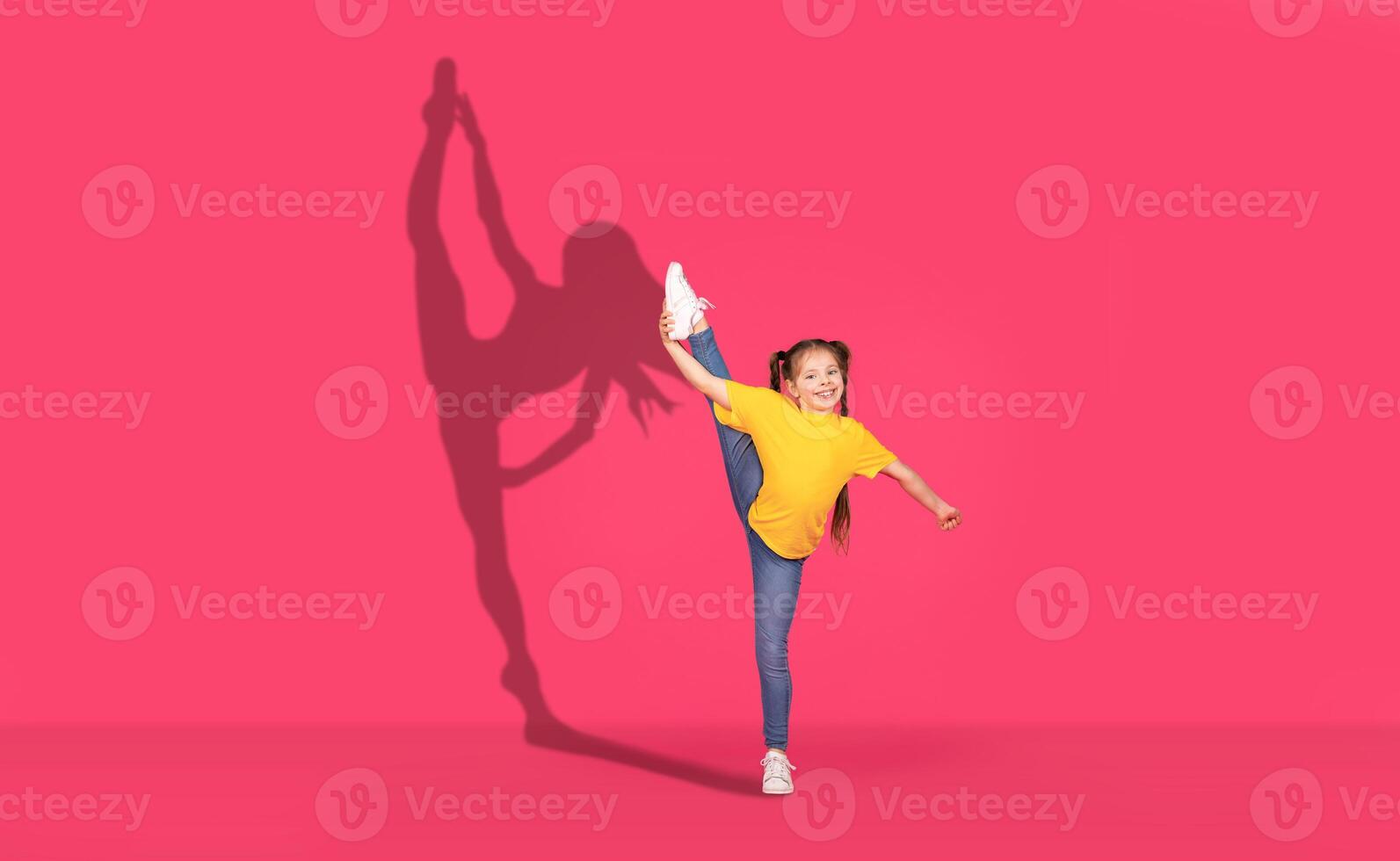 un' gioioso giovane ragazza nel un' giallo camicia e jeans esegue un' alto gamba calcio con entusiasmo foto