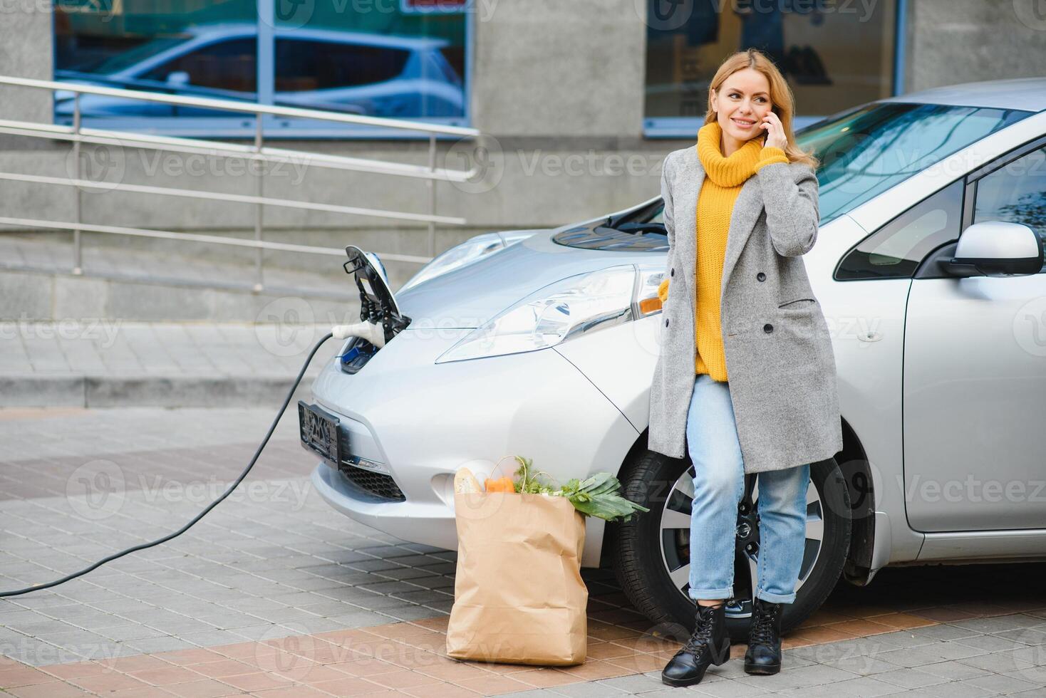 avendo conversazione di utilizzando Telefono. donna su il elettrico macchine caricare stazione a giorno. marca nuovo veicolo foto