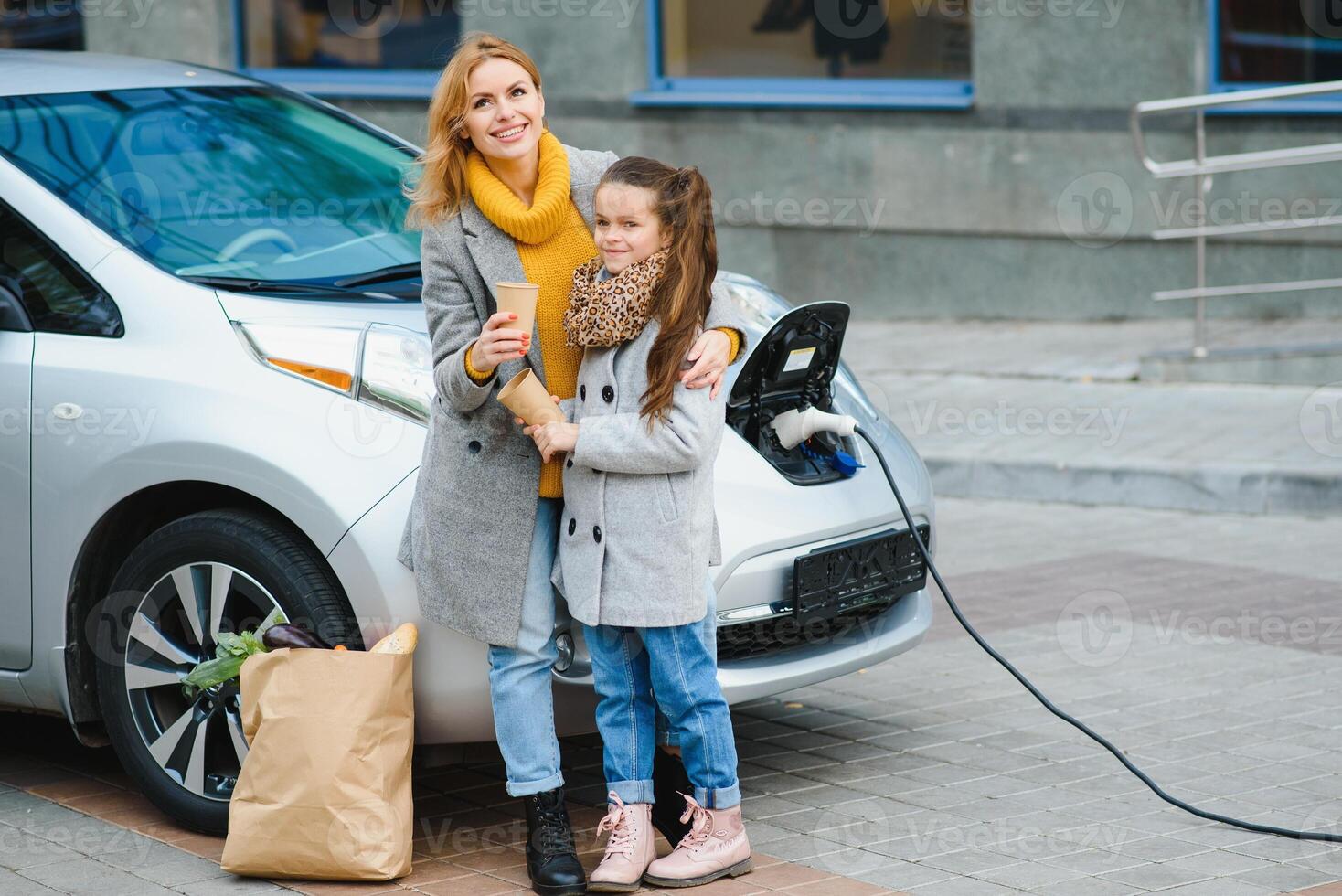 madre con figlia ricarica elettro auto a il elettrico gas stazione e parlare su mobile Telefono foto