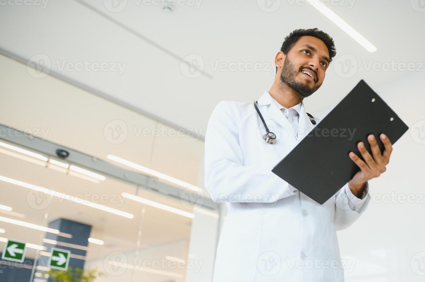 ritratto di contento amichevole maschio indiano latino medico medico lavoratore indossare bianca cappotto con stetoscopio in giro collo in piedi nel moderno privato clinica guardare a telecamera. medico assistenza sanitaria concetto foto