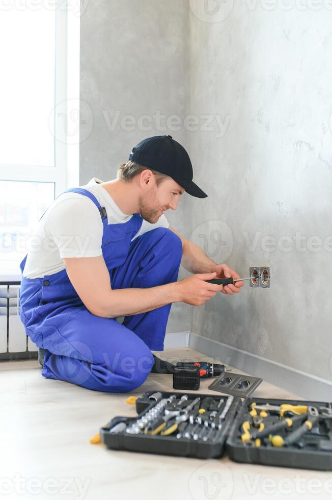 fiducioso professionale elettricista nel uniforme utilizzando Cacciavite mentre sostituzione un' presa di corrente nel appartamento dopo rinnovamento opera foto