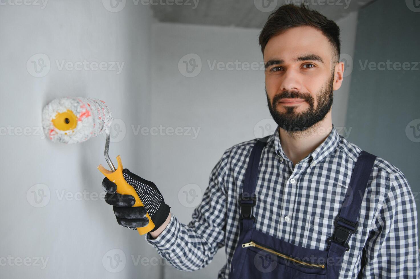 giovane sorridente professionale lavoratore nel blu uniforme in piedi con dipingere rullo nel nuovo appartamento per riparazione al di sopra di grigio muri sfondo, copia spazio. foto