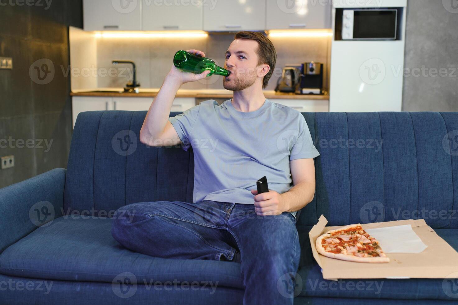 contento giovane uomo potabile birra e mangiare Pizza quando Guardando gioco su tv a casa foto