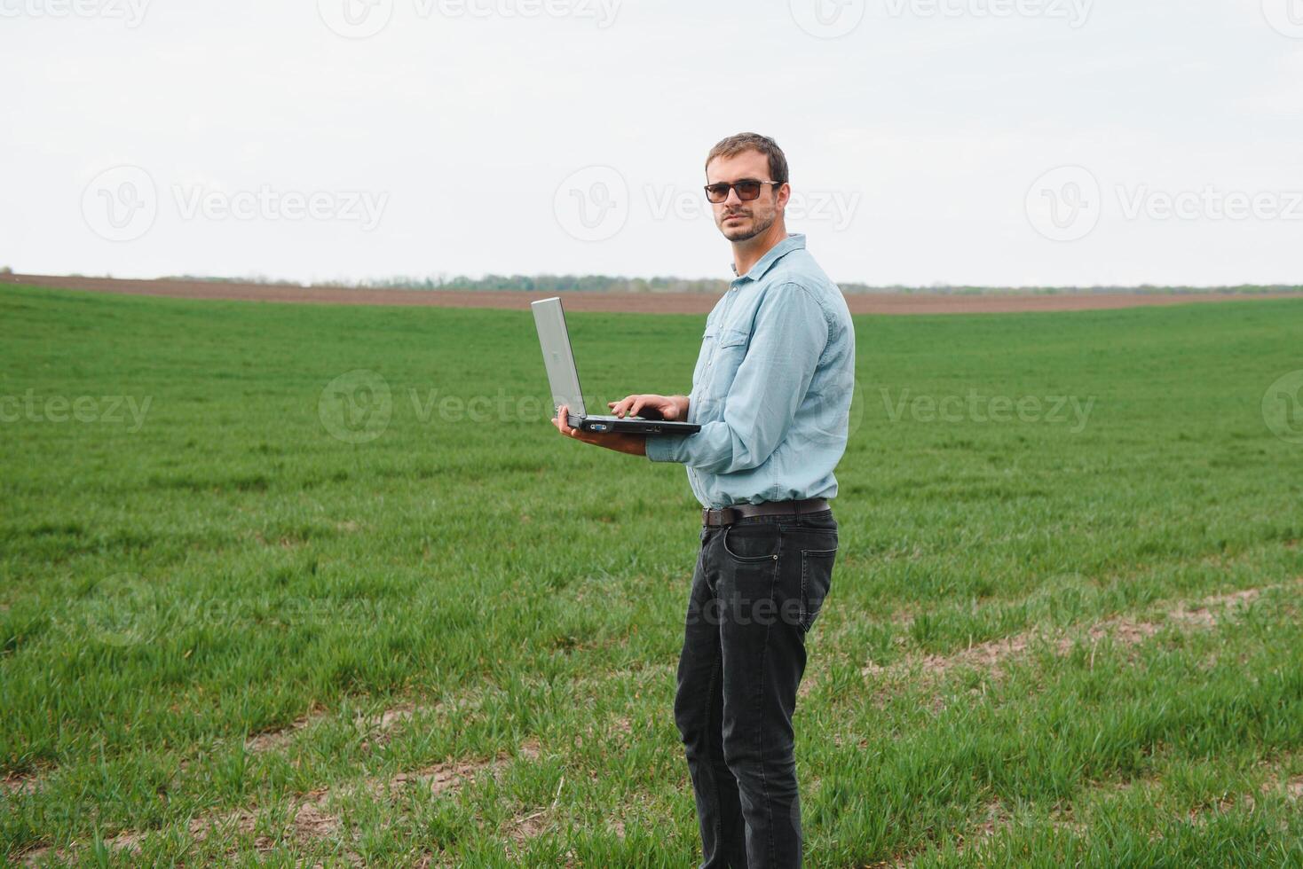 contadino in piedi nel giovane Grano campo l'esame Ritaglia e guardare a il computer portatile. foto