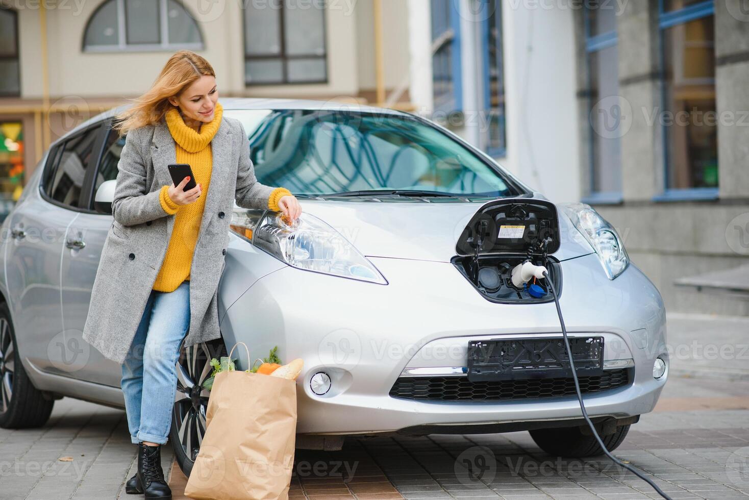 utilizzando smartphone mentre in attesa. donna su il elettrico macchine caricare stazione a giorno. marca nuovo veicolo foto