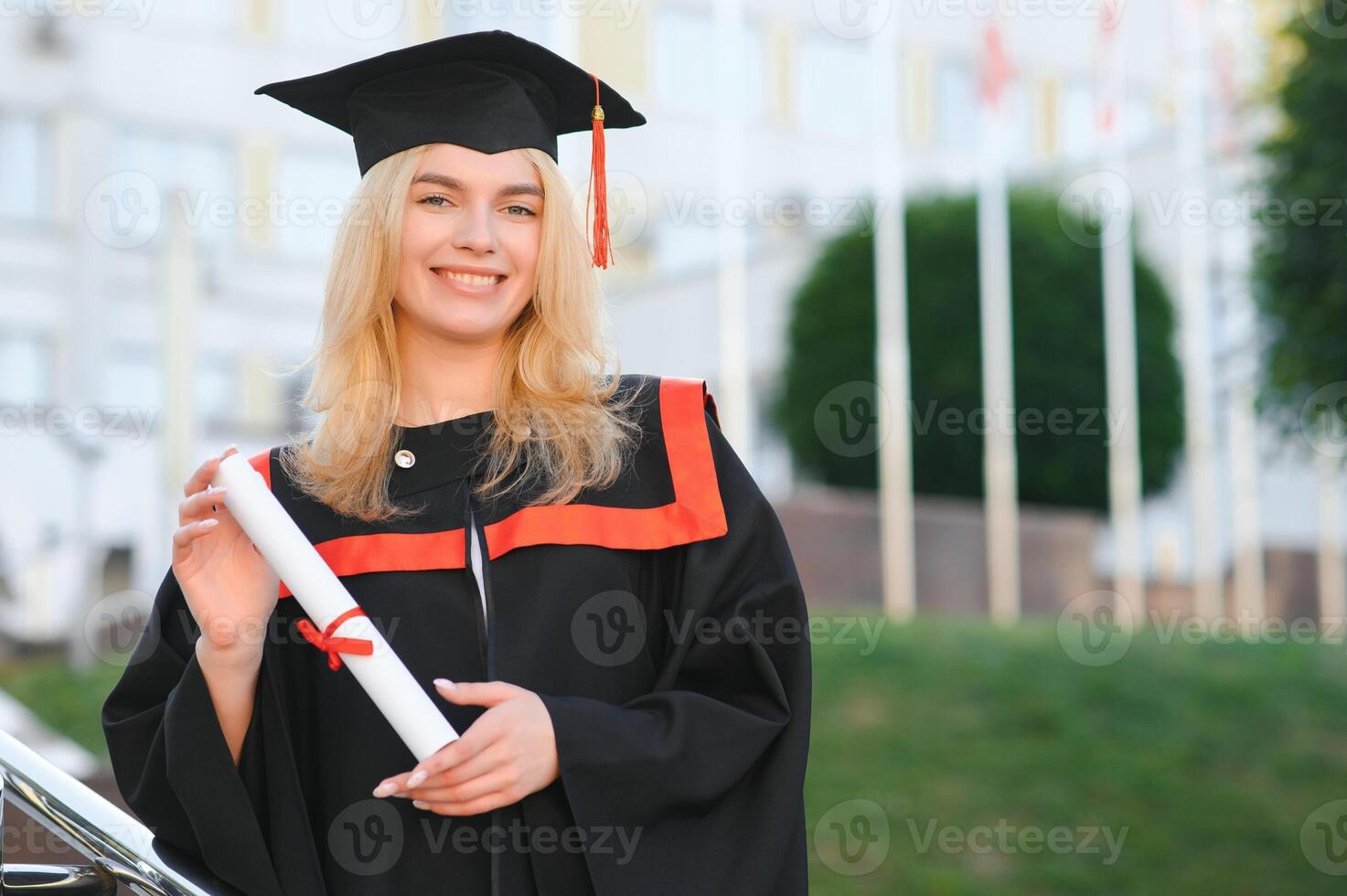 contento carino caucasico grad ragazza è sorridente. lei è nel un' nero mortaio asse, con rosso nappa, nel abito, con simpatico Marrone Riccio capelli, diploma nel mano foto