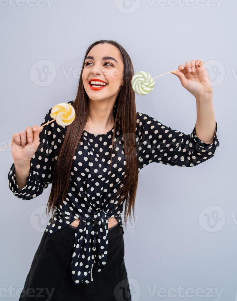 giovane bellezza nel tratteggiata camicia con Due Lecca-lecca nel mani. allegro signora con caramelle al di sopra di grigio sfondo. foto