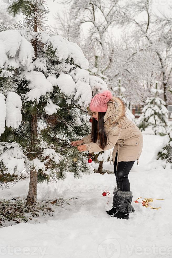 bella donna in abiti invernali caldi che decorano l'albero di natale in un parco in una giornata nevosa foto