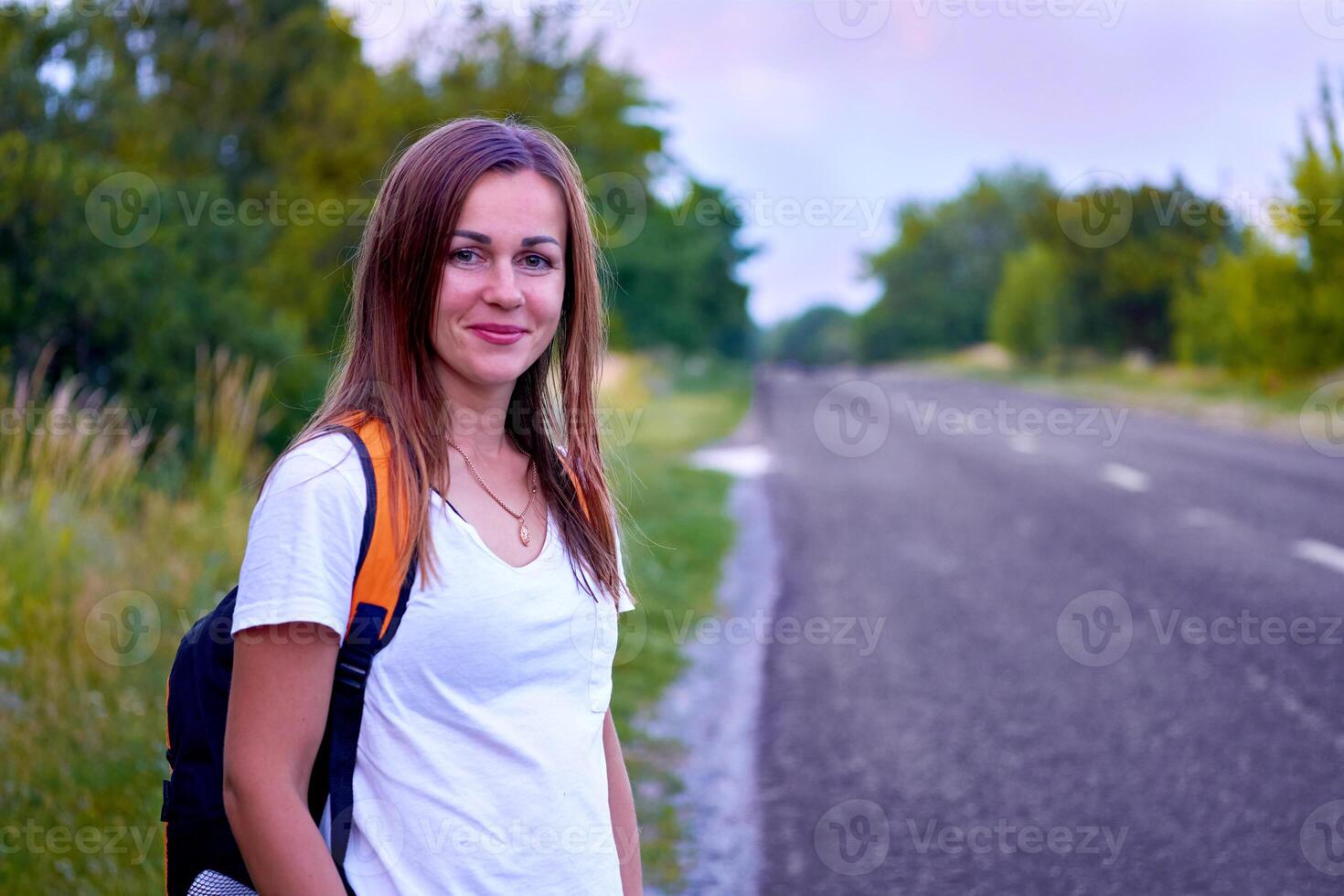 giovane in viaggio allegro ragazza sta in attesa di il asfalto strada con alberi foto