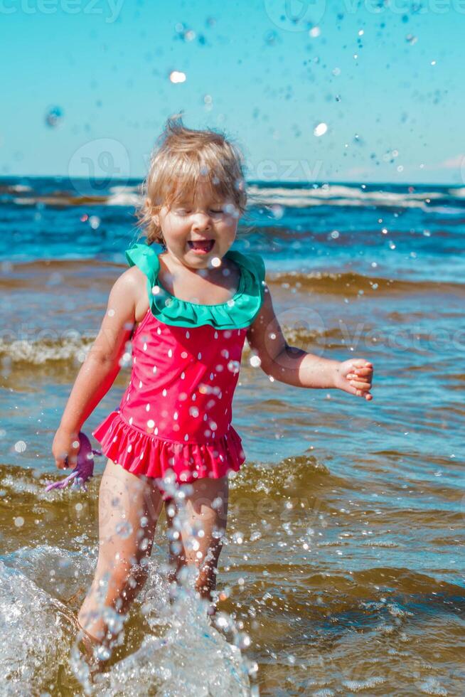 giovane contento bambino ragazza di europeo aspetto età di 4 avendo divertimento nel acqua su il spiaggia e spruzzi,tropicale estate vocazioni,vacanze.a bambino gode il mare.verticale foto. foto