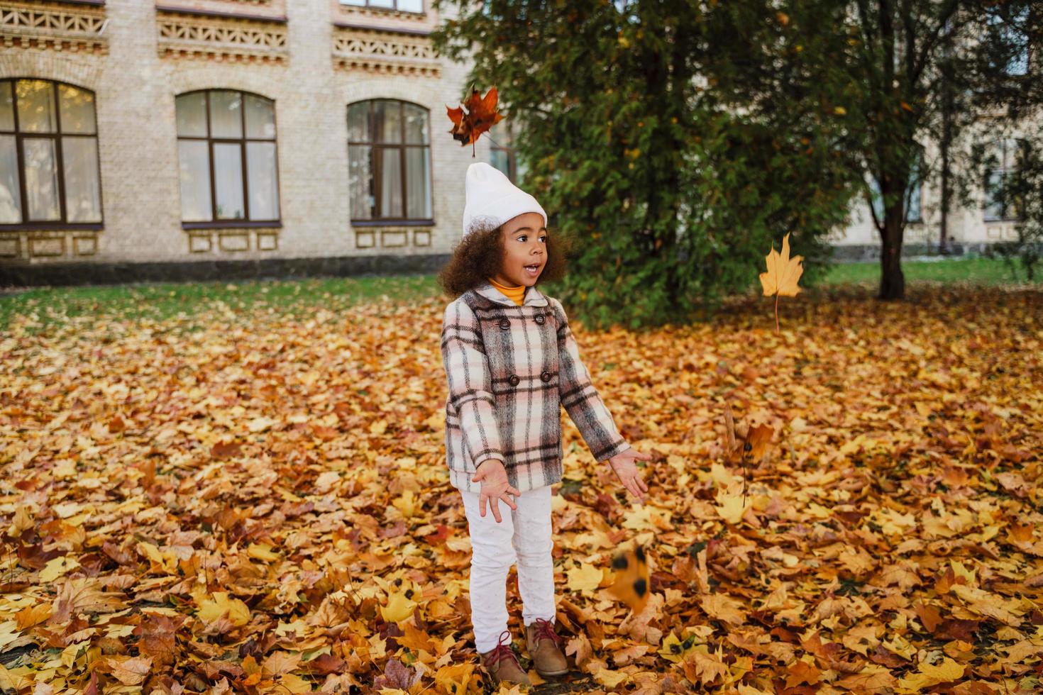 ragazza nera che indossa un cappotto che si diverte con le foglie cadute nel parco autunnale foto