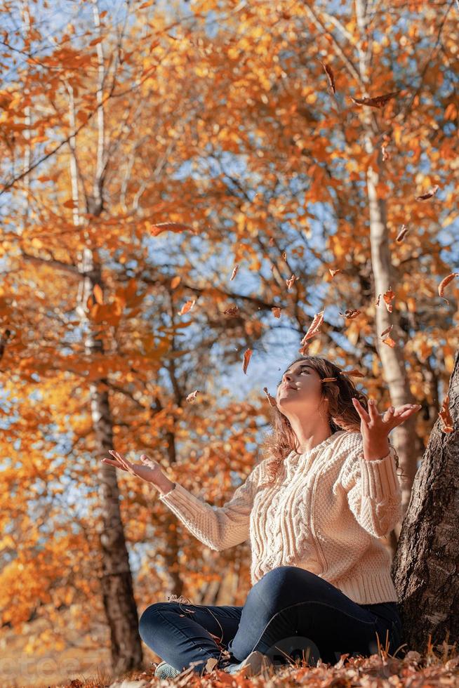 giovane donna che lancia foglie gialle in aria seduta nella foresta autunnale foto