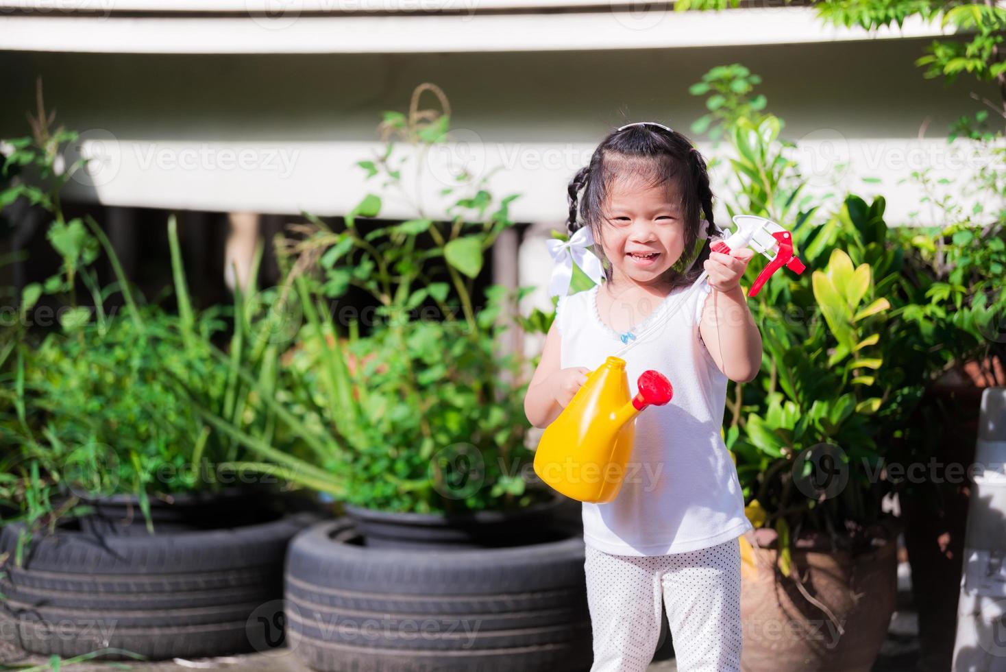 ragazza carina teneva in mano un annaffiatoio giallo, sorridendo dolcemente nel giardino di un piccolo albero davanti alla casa. i bambini aiutano nelle faccende domestiche. ugello spray per bambini. serate estive o primaverili. Bambino di 3 anni. foto