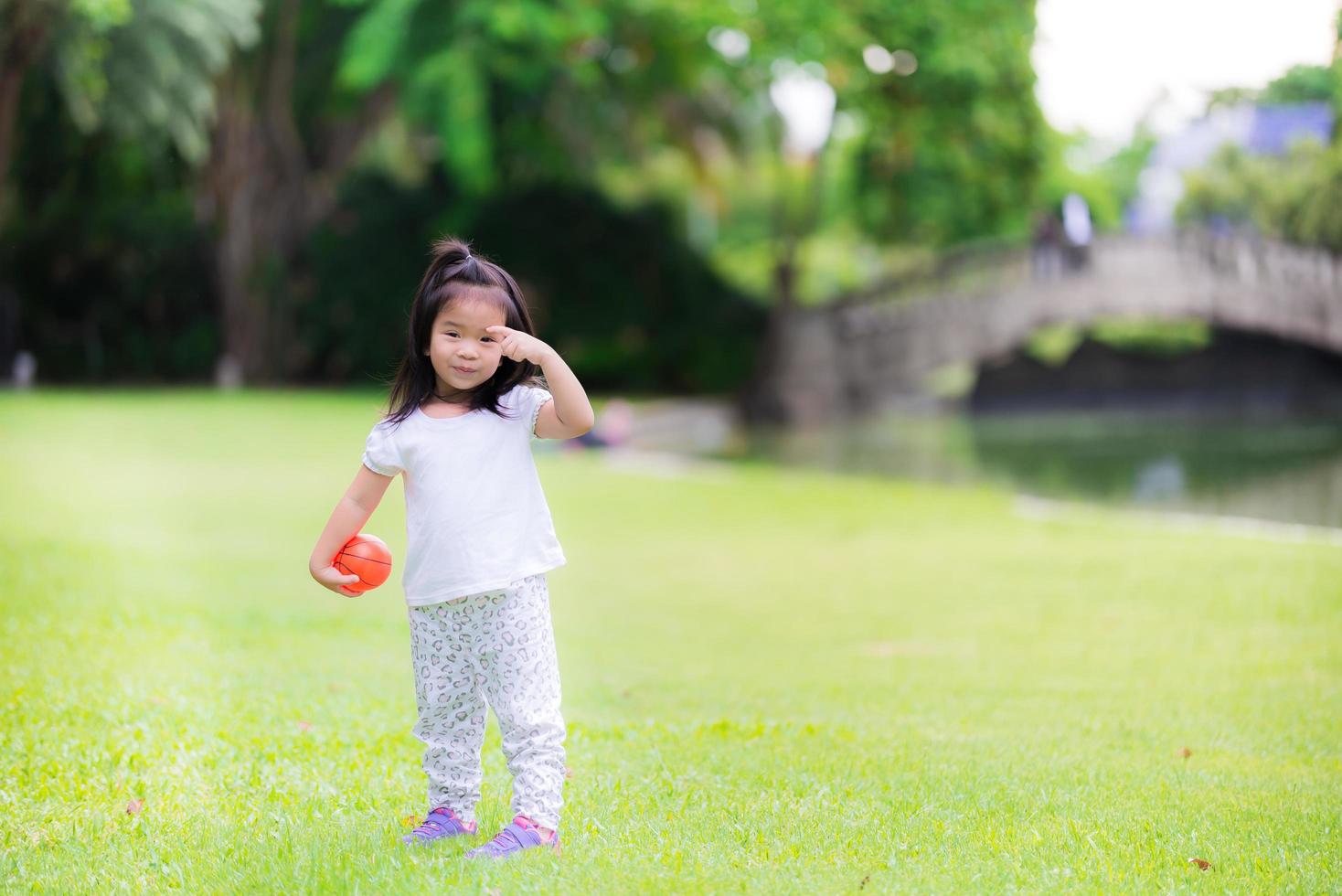 ragazza felice che gioca piccola palla arancione in erba verde con la famiglia. picnic con i genitori in vacanza. inverno nel centro della Thailandia. clima fresco. carino bambino di 3 anni, sorriso dolce, bambino che si gratta le sopracciglia foto