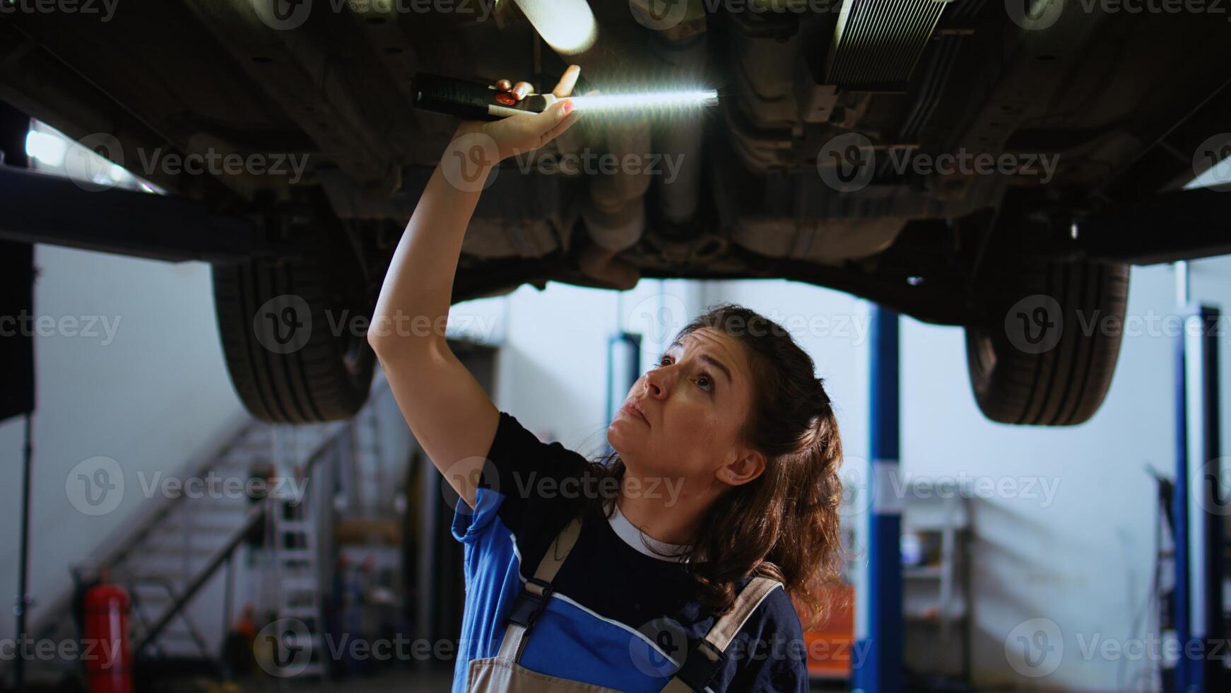 concesso in licenza meccanico Lavorando su sospeso auto nel box auto, controllo componenti durante routine Manutenzione. auto riparazione negozio dipendente a piedi sotto veicolo, ispezionando esso utilizzando opera leggero foto