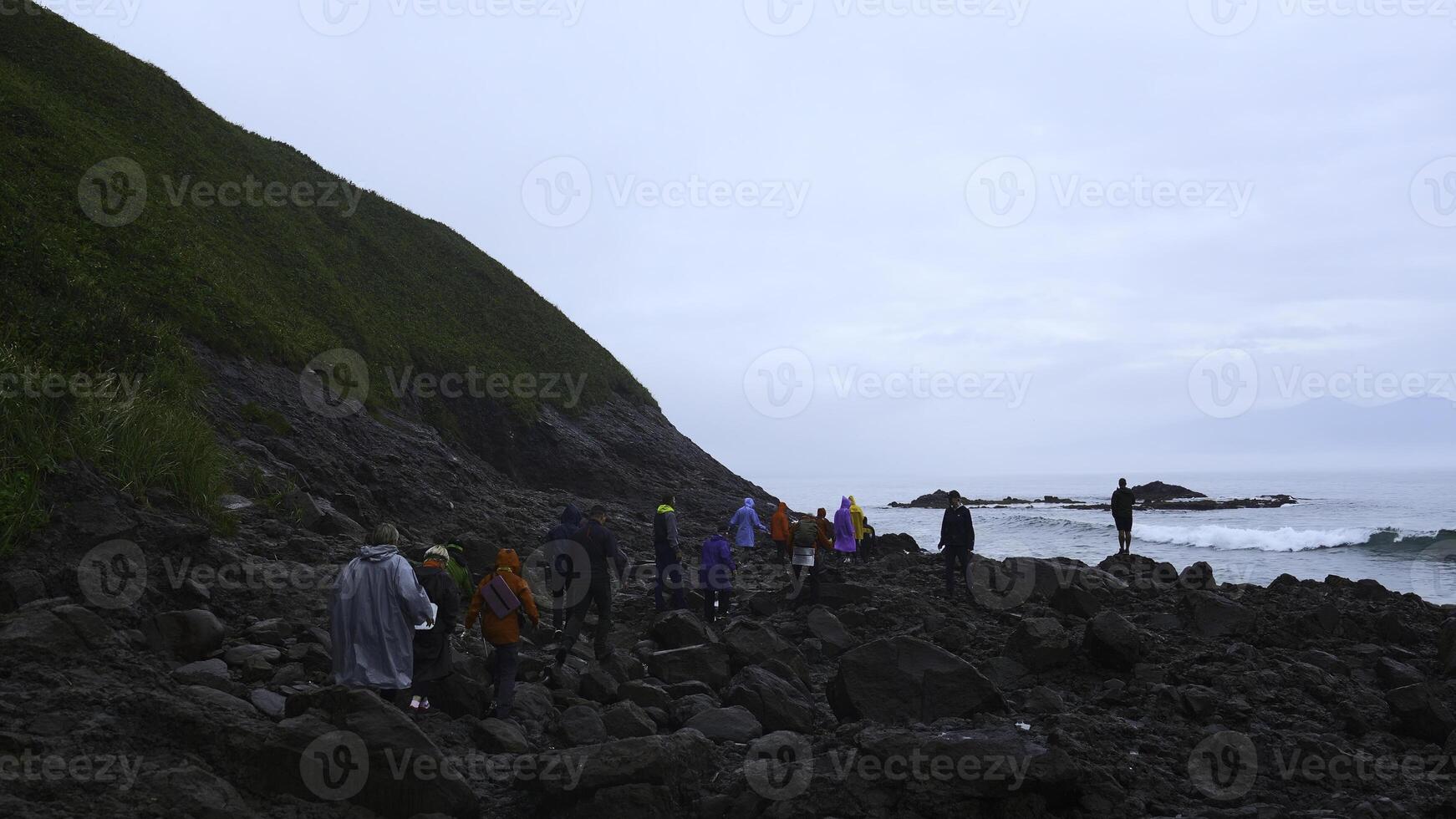 gruppo di turisti passeggiate su pietra costa. clip. turisti camminare su pericoloso pietra riva nel nuvoloso tempo atmosferico. gruppo di turisti su costa con nero rocce di settentrionale paesaggio foto