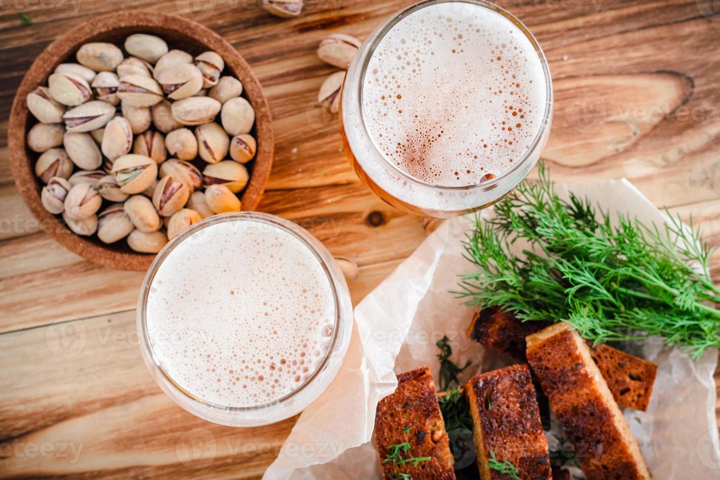 due bicchieri di birra, toast con crosta, aneto e pistacchi su un tavolo di legno. messa a fuoco selettiva sulla schiuma foto