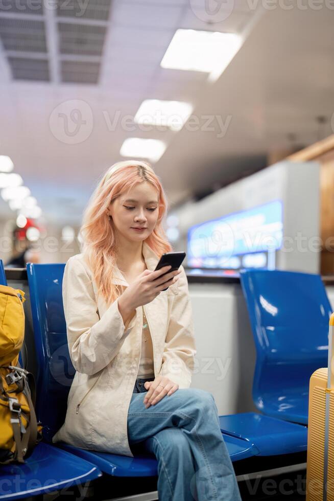 contento asiatico turista donna utilizzando mobile smartphone con valigia in viaggio fra aspetta per volo nel aeroporto terminale, volo dai un'occhiata in, turista viaggio viaggio concetto foto