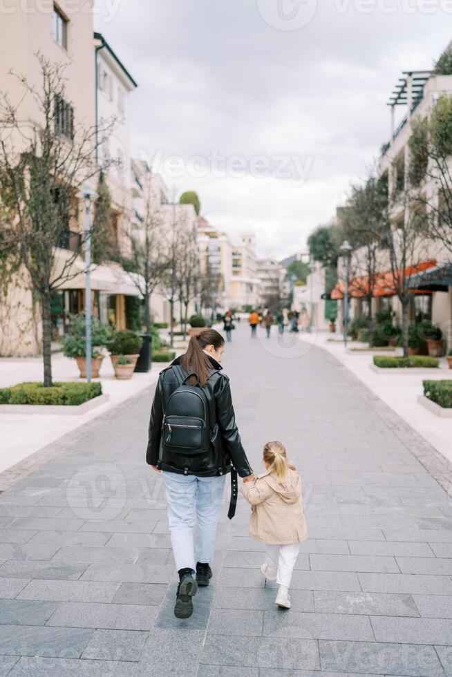 madre e un' poco ragazza camminare Tenere mani lungo il strada passato palazzi. indietro Visualizza foto