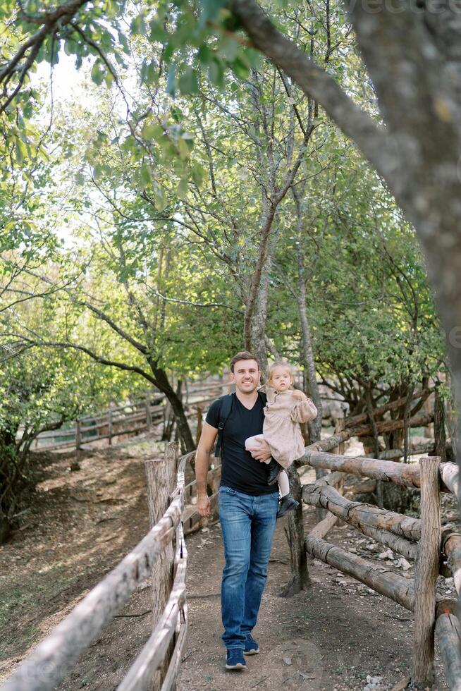 papà con un' poco ragazza nel il suo braccia passeggiate lungo un' sentiero fra recinzioni nel il parco foto