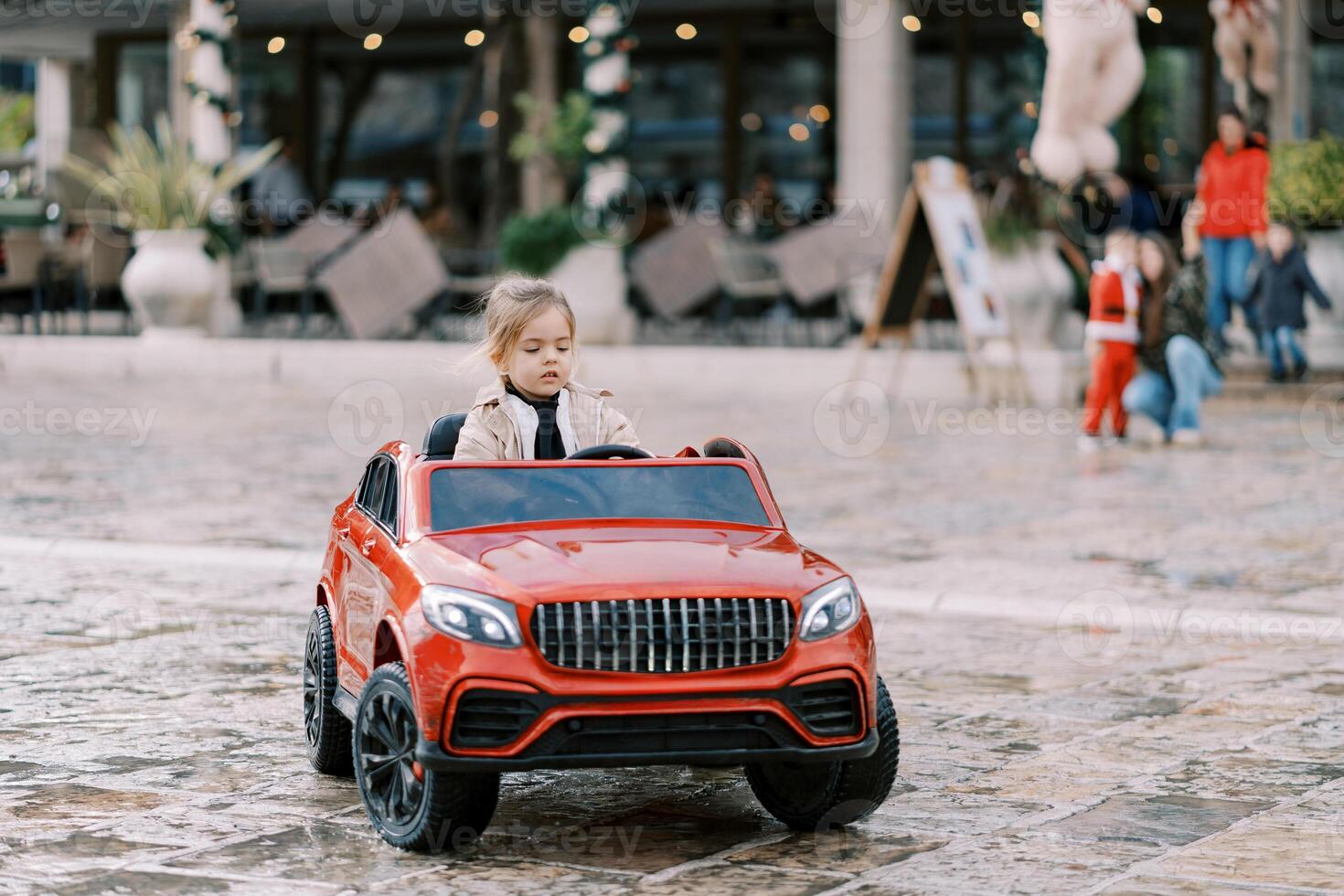 poco ragazza unità un' rosso giocattolo auto attraverso il piazza foto