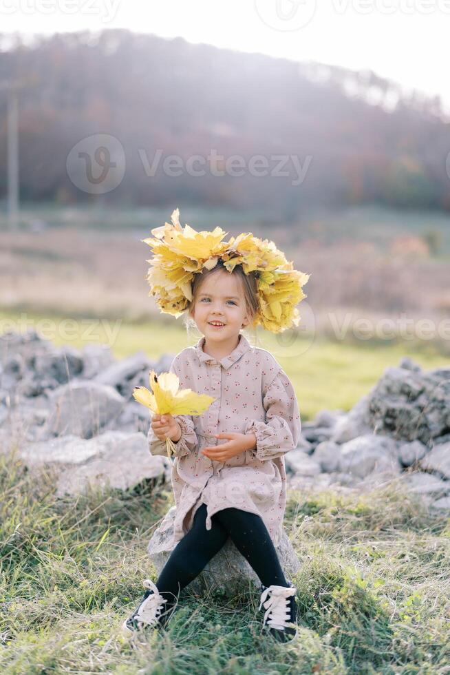 poco sorridente ragazza nel un' ghirlanda di autunno le foglie si siede su un' pietra su il prato con giallo le foglie nel sua mano foto