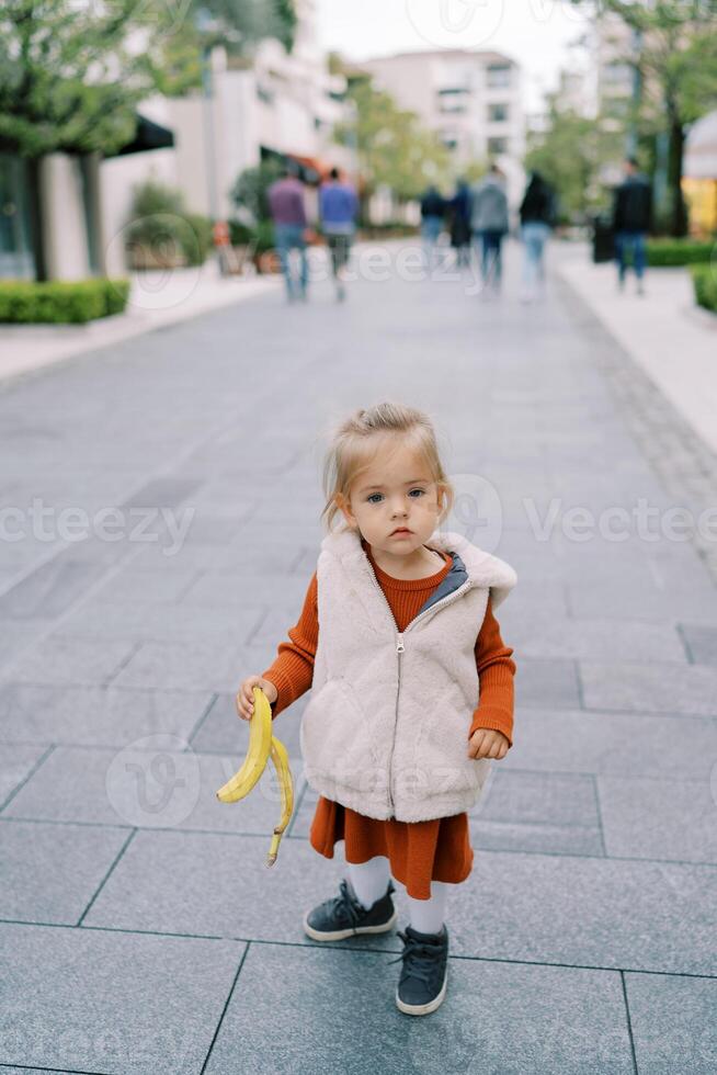 poco ragazza trasporta un' Banana pelle giù il strada foto