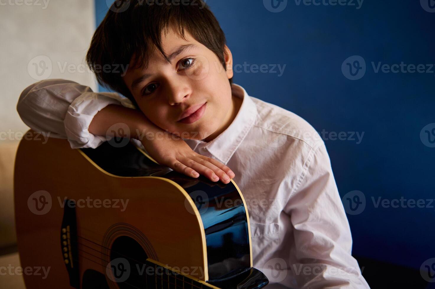 autentico ritratto di un' caucasico carino adolescenziale ragazzo nel bianca casuale camicia, in posa con il suo acustico classico chitarra, sorridente guardare a telecamera, seduta nel il musica studio al di sopra di blu parete sfondo foto