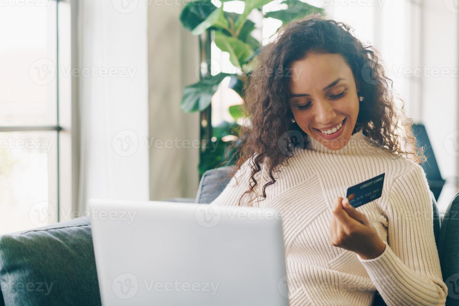 donna latina che utilizza laptop e mano che tiene la carta di credito per lo shopping sul divano foto