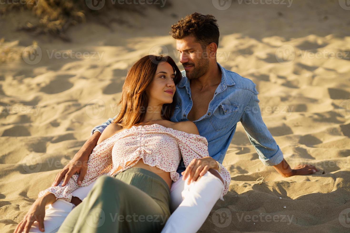 coppia attraente che si siede sulla sabbia della spiaggia che si gode l'un l'altro foto