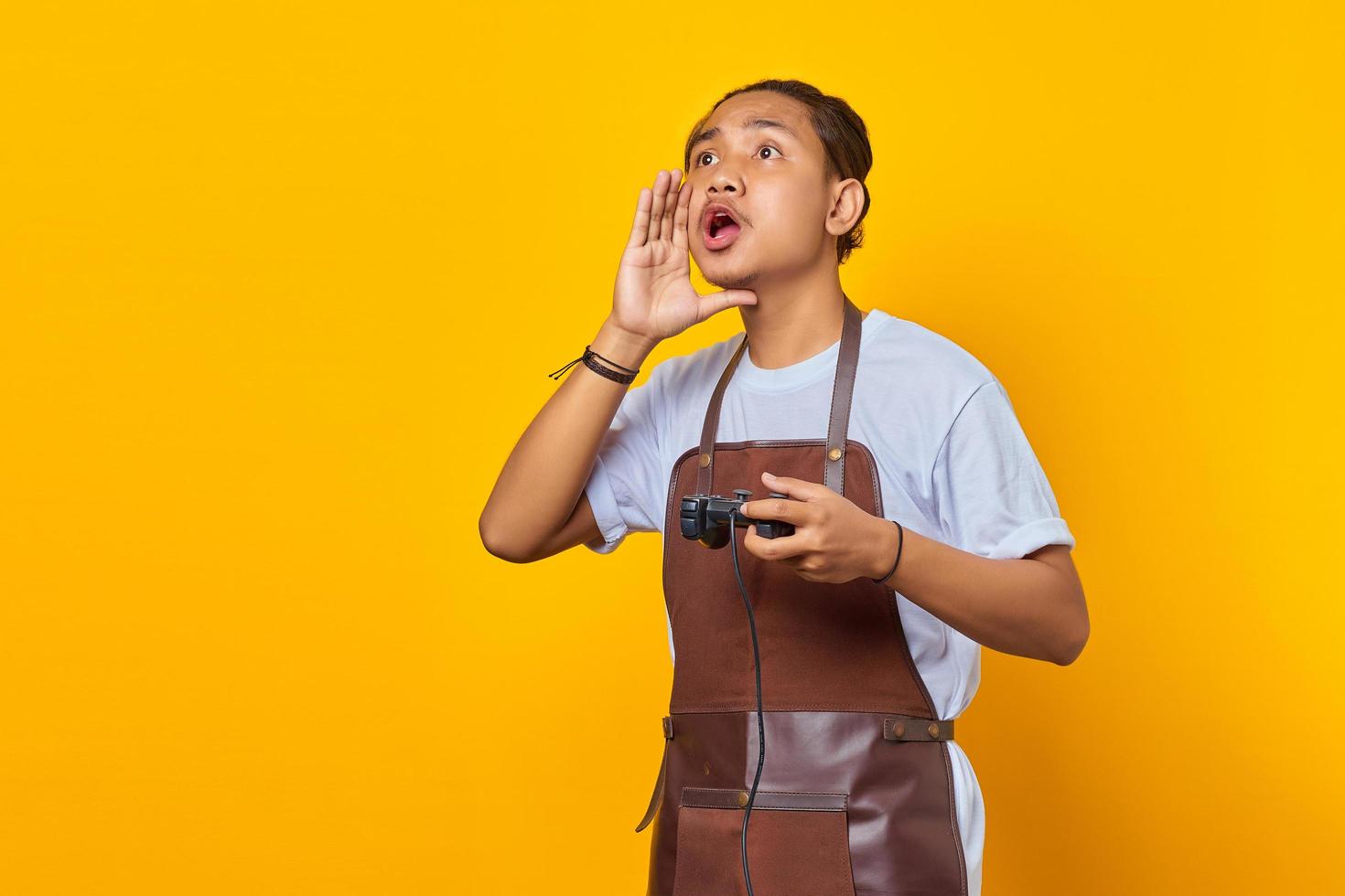 ritratto di un giovane asiatico che indossa un grembiule che tiene in mano un controller di gioco con un grido a bocca aperta che incoraggia isolato su sfondo giallo foto