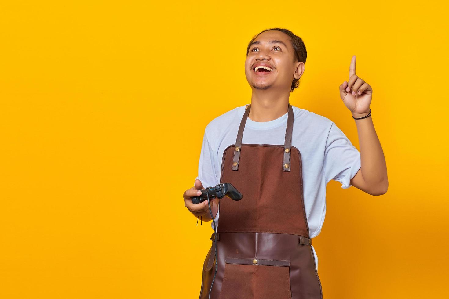 giovane uomo che indossa un grembiule che tiene il controller di gioco sorridendo allegramente mentre punta il dito su sfondo giallo foto