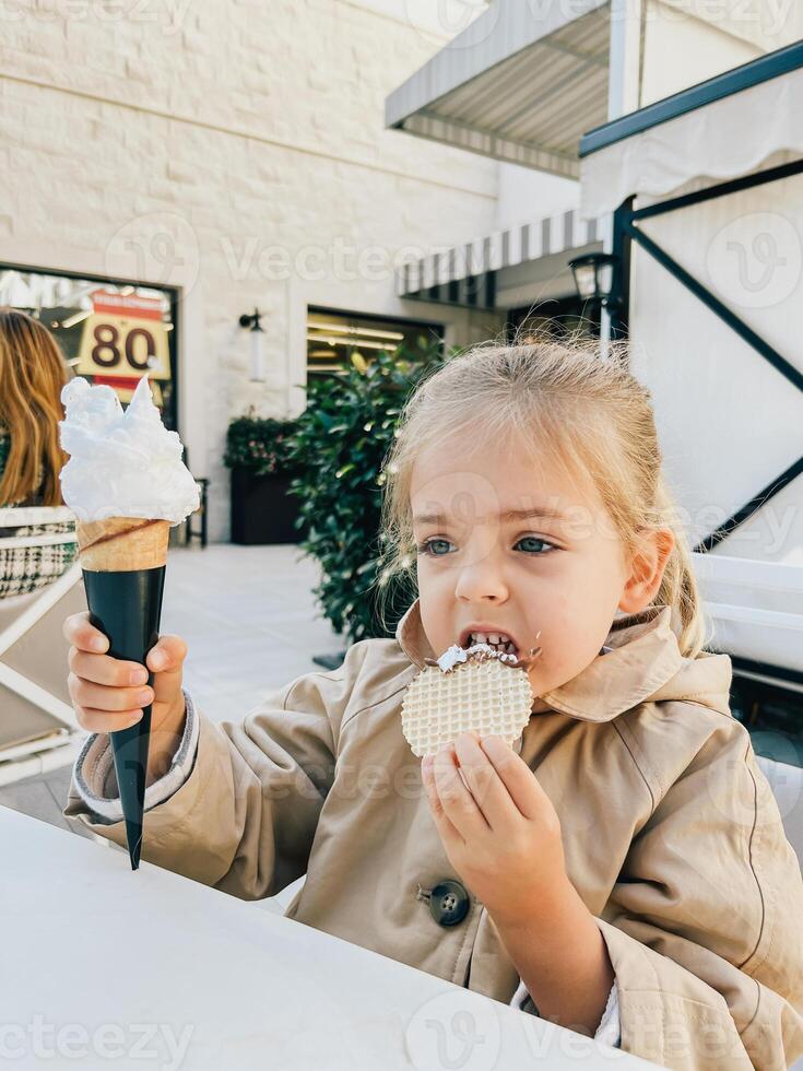 poco ragazza mangia un' cialda a partire dal un ghiaccio crema cono mentre seduta a il tavolo foto