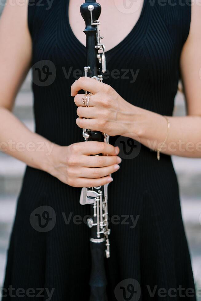 musicista ragazza nel un' nero vestito detiene un' clarinetto nel sua mani nel davanti di sua il petto. ritagliato. senza volto foto