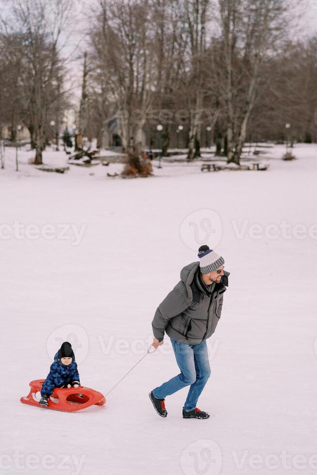 sorridente papà trasporta un' poco ragazzo su un' slitta attraverso un' nevoso pianura a il bordo di un' foresta foto