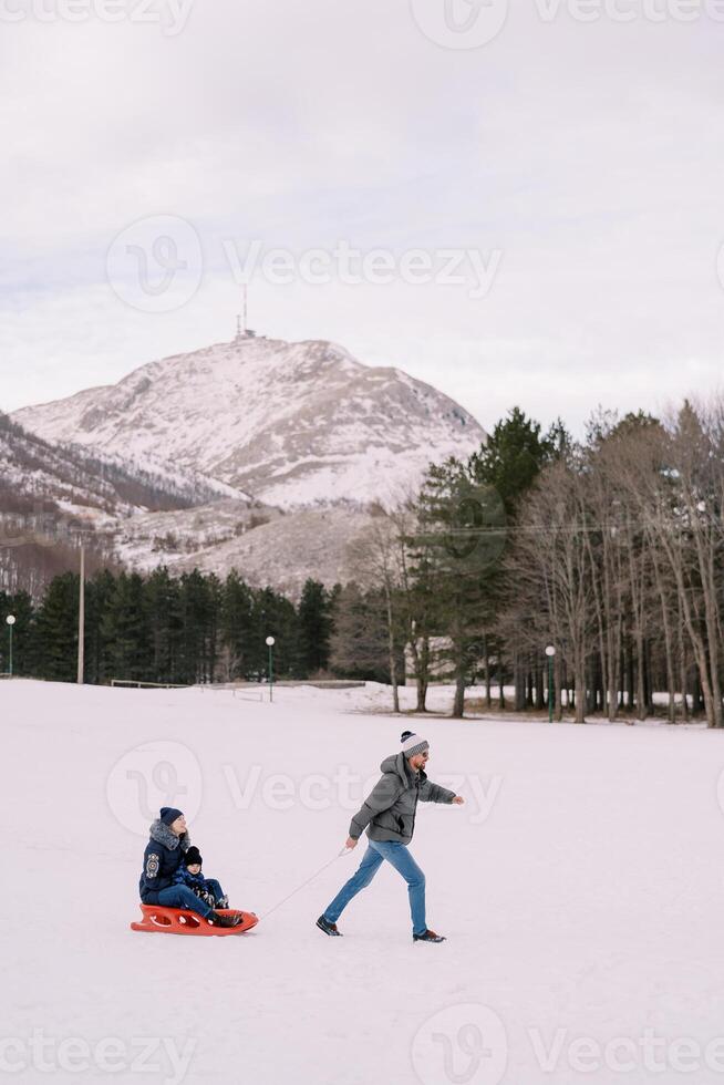 papà trasporta mamma e bambino su un' slitta attraverso un' nevoso pianura a il piede di il montagne foto