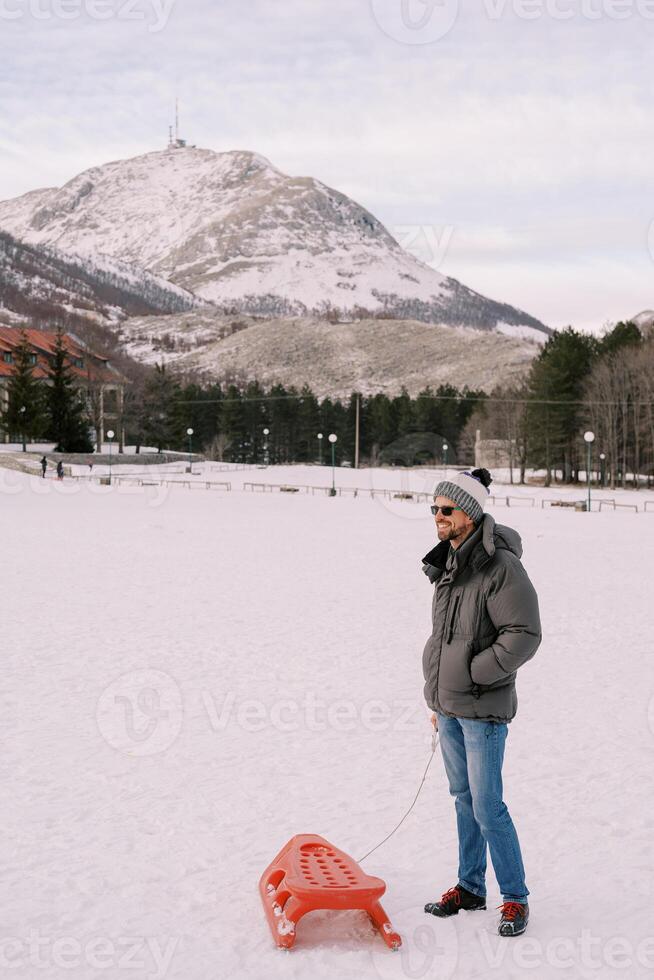 sorridente giovane uomo nel occhiali da sole in piedi su nevoso pianura Tenere slitta di corda con mani nel tasche foto