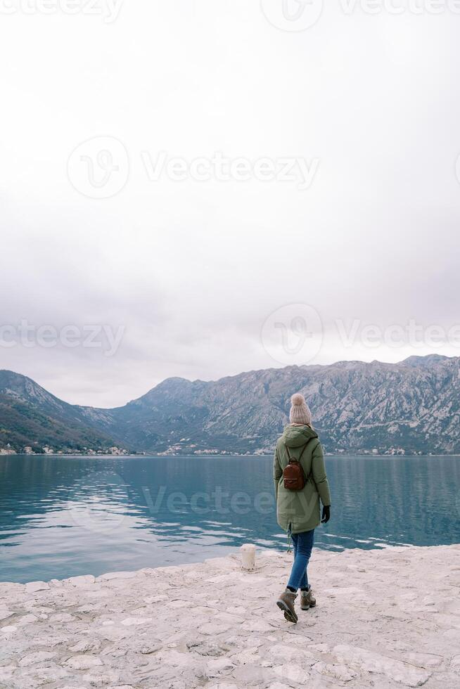 donna con un' zaino passeggiate lungo il molo di il mare e sembra a il montagne. indietro Visualizza foto