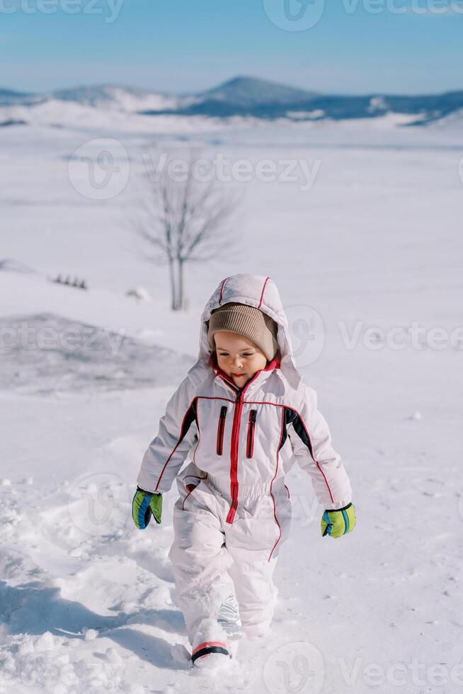 poco ragazza passeggiate attraverso in profondità cumuli di neve nel un' radura, guardare a sua piedi foto